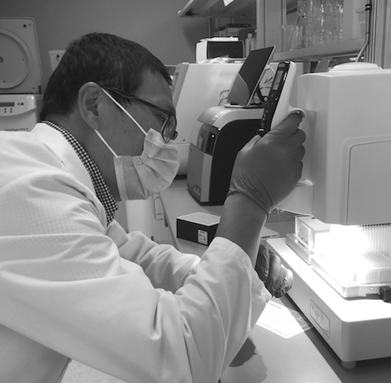 Black and white image of a lab scientist in a mask and gloves handling a micropipette.